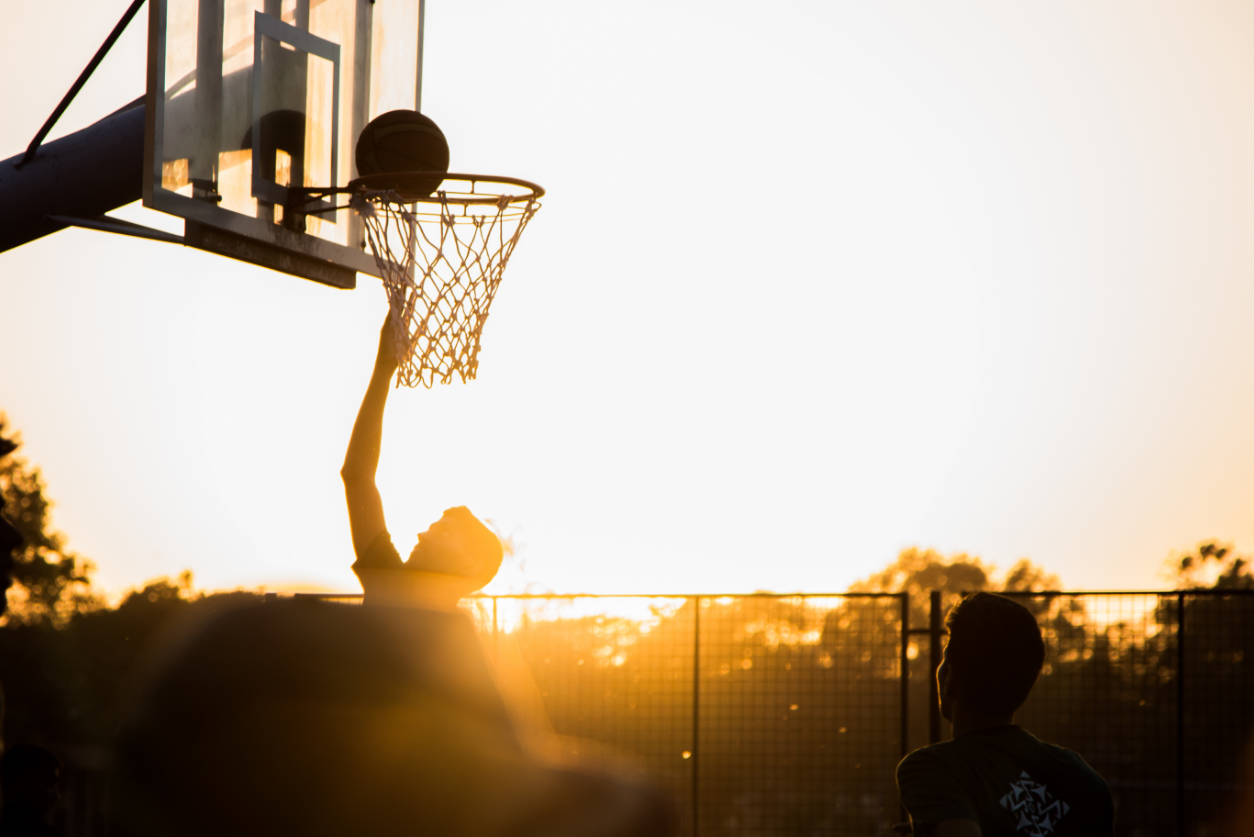 A teenage boy making a layup to a basketball hoop on an outside court at sunset