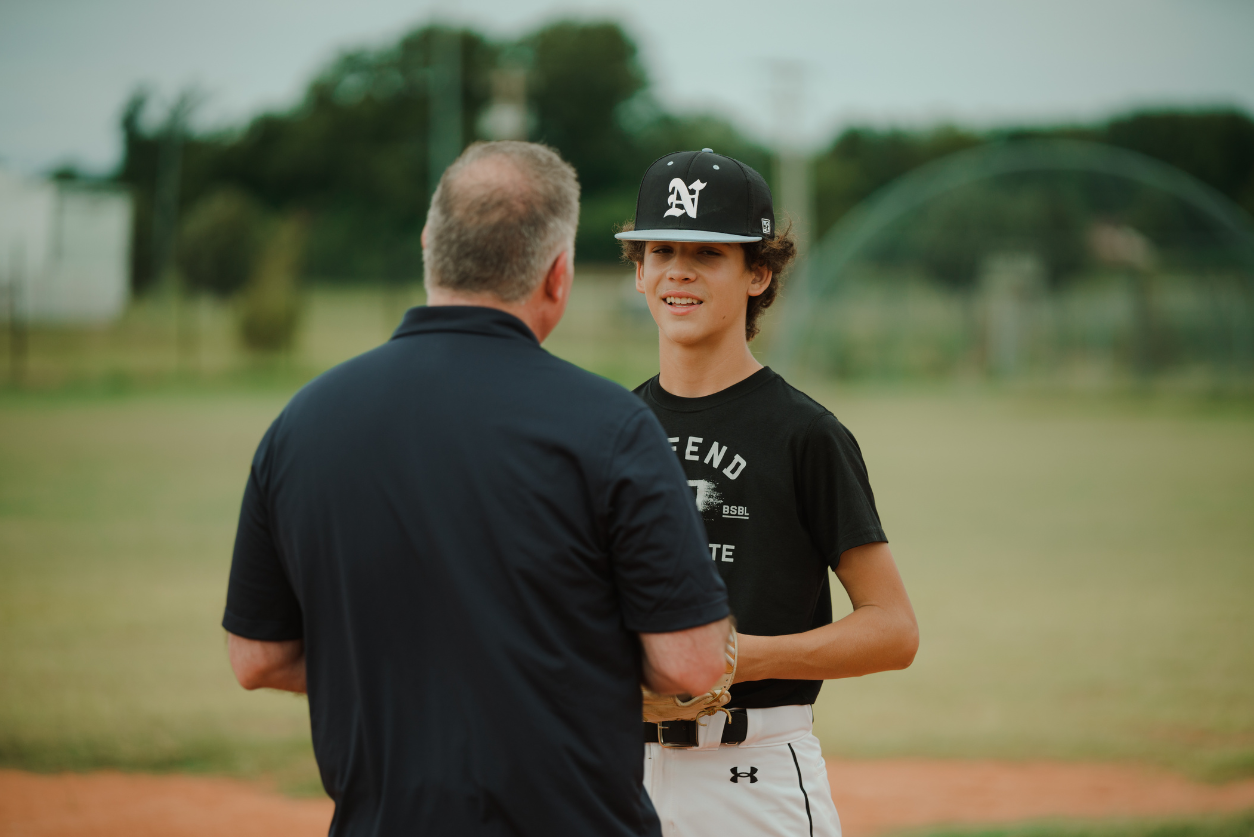 A sports psychologist talks with a baseball player in uniform on the field.