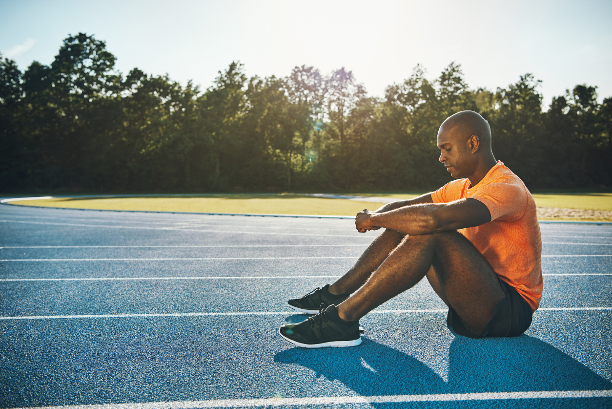 Male athlete in an orange shirt sits on the track alone with his knees up, and his arms resting on his knees.