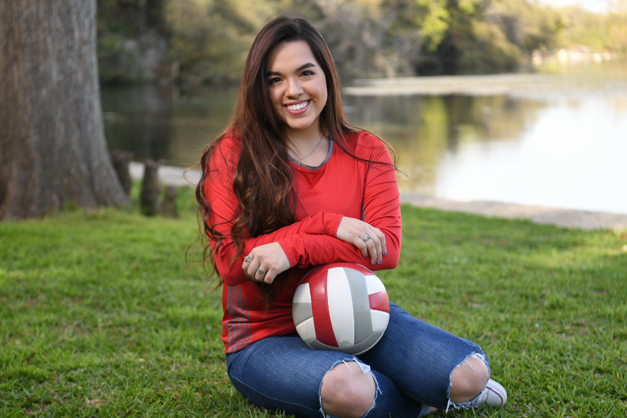 A young woman with dark brown hair volley ball player, wearing jeans ripped in the knees and a long sleeved red shirt, sits on the grass holding a volley ball.