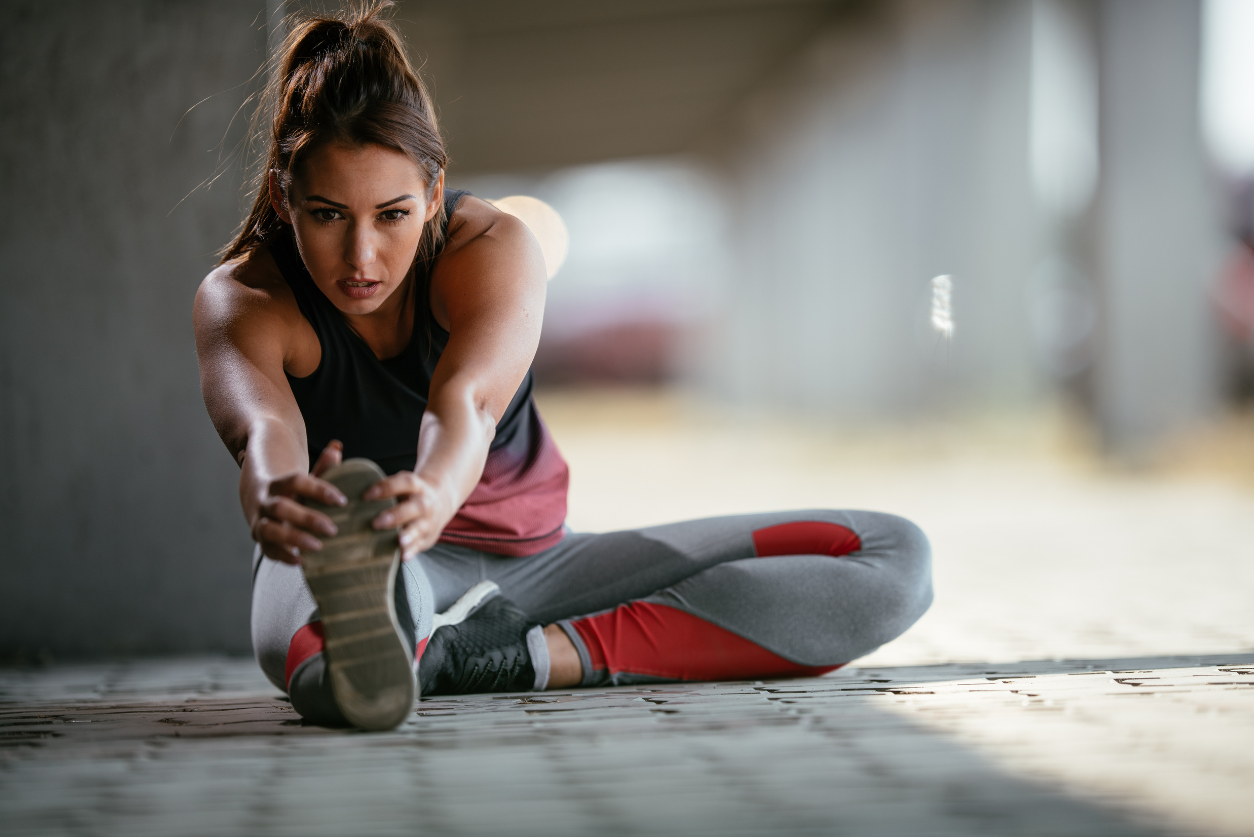 A female athlete with long, dark hair in a ponytails sits on the floor stretching her legs and looks focused.