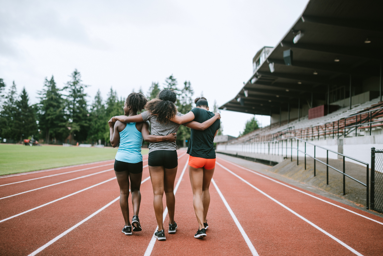 Three black female athletes walk down the outdoor track with arms around each other.