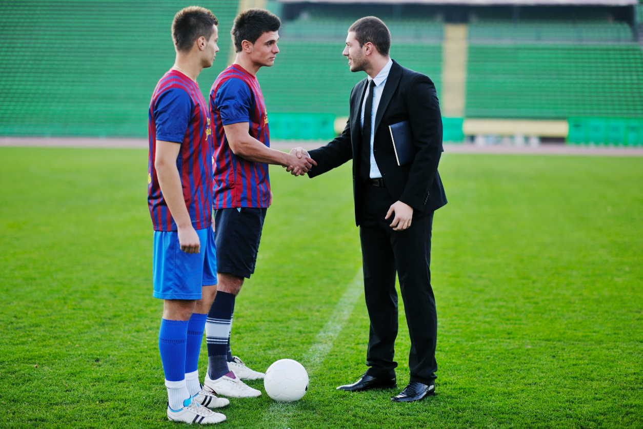 A man in a suit stands on a soccer field shaking hands with a young soccer player in uniform.