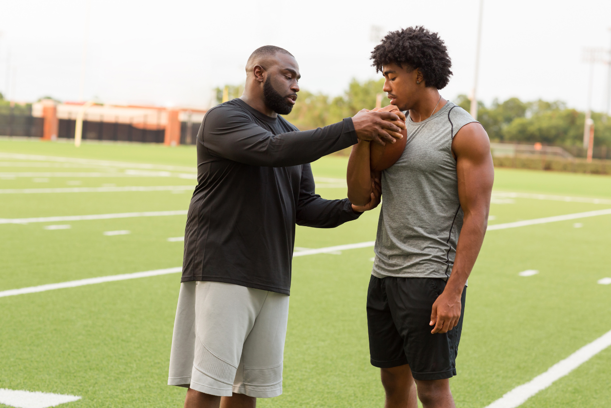 An adult male coach teaches a young male athlete how to carry a football.