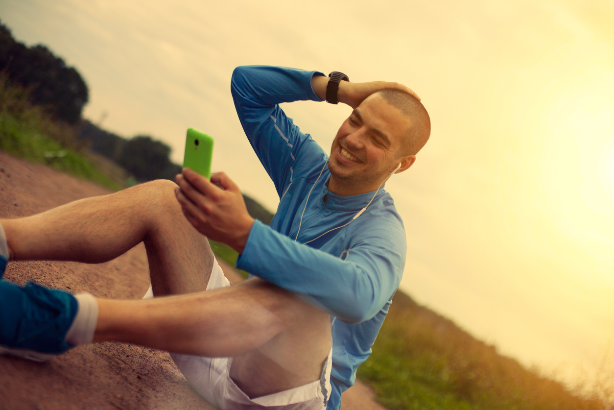 A young man sitting on the ground wearing running clothes smiles while he looks at his phone.