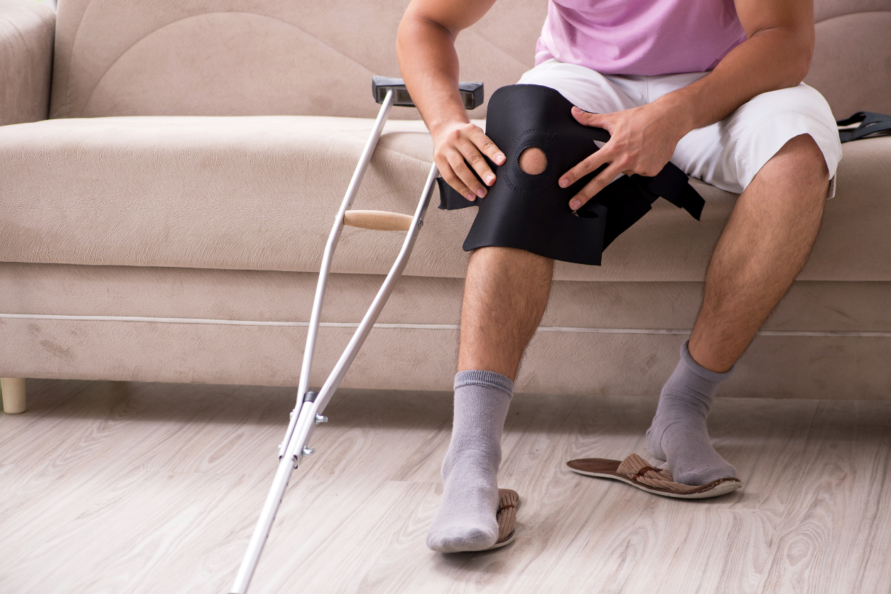 A male wearing white shorts adjusts a knee brace while sitting on a couch with crutches leaning against it.