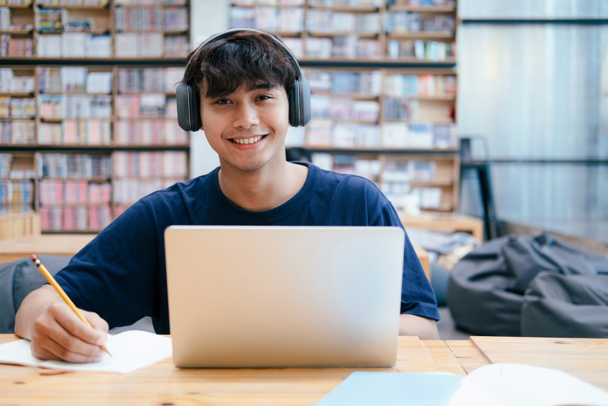 A young male smiles at the camera wearing headphones, sitting in front of a laptop in a library.