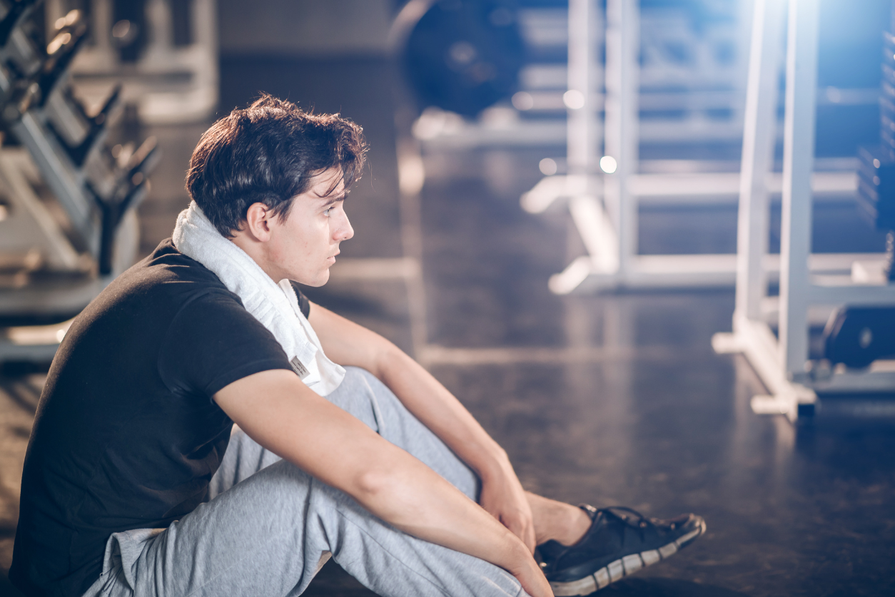 A young man sits on the gym floor with a white towel around his neck and his arms propped up on his knees.