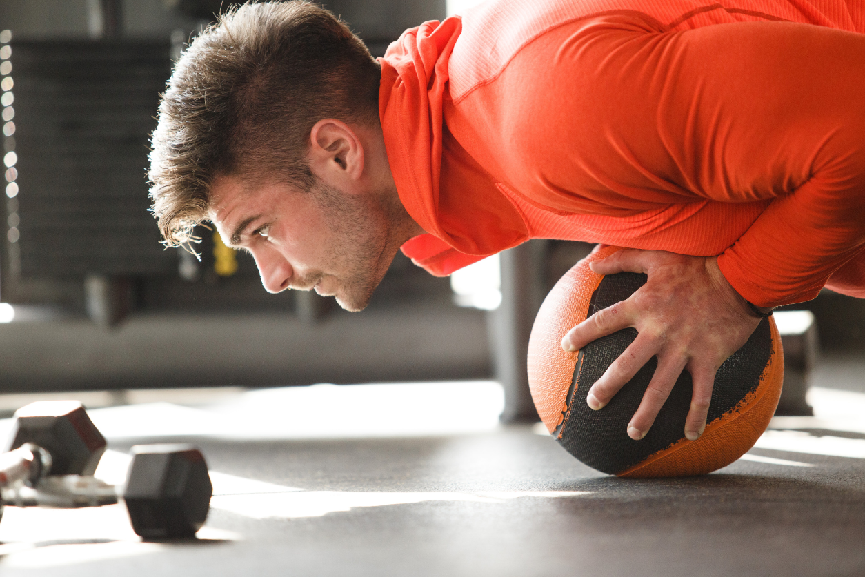 A male athlete wearing a long sleeve orange shirt balances his upper body with his hands on a medicine ball looking focused.