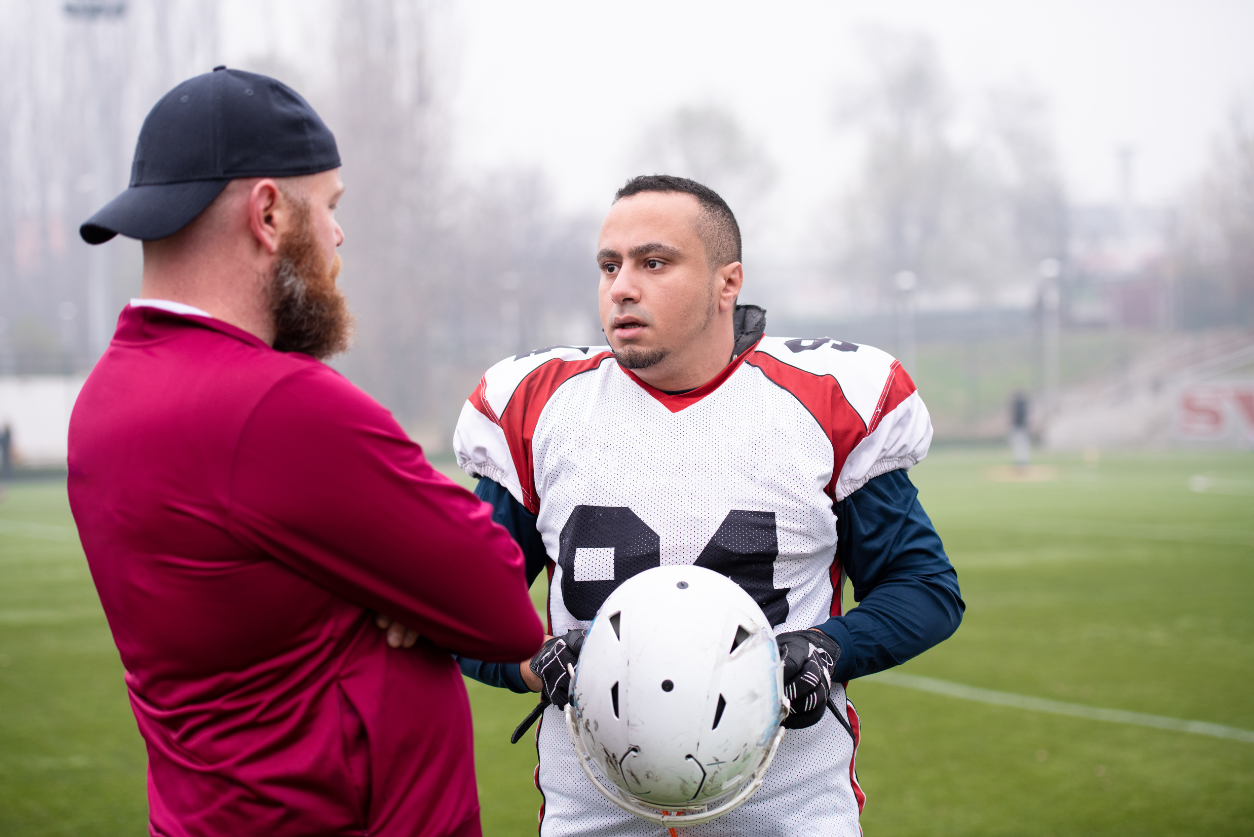 A coach in a red shirt stands with arms crossed on the field talking with a player in a white jersey holding his white helmet.