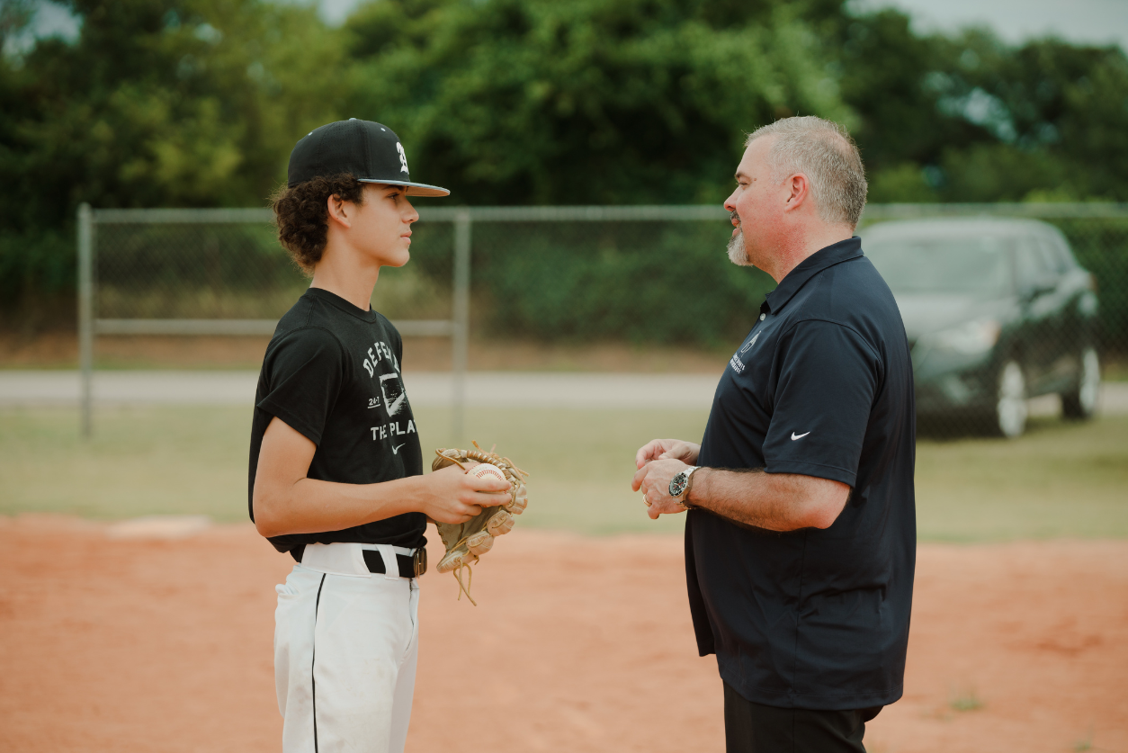 A baseball player talks on the field with a coach .