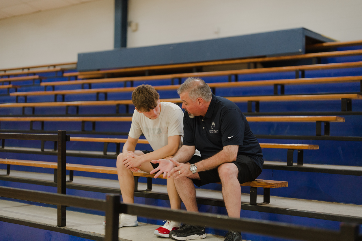 A man in a dark blue polo sits on bleachers and talks with a male basketball player in a white jersey.