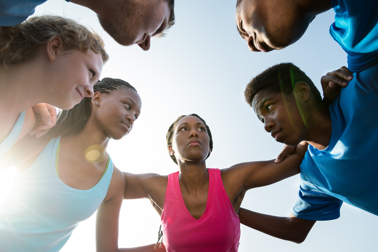 A group of young athletes huddles with arms on their shoulders with the sun shining through.